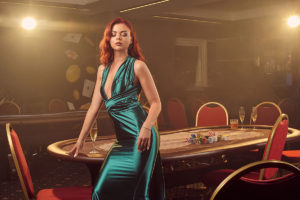 alluring-redheaded-maiden-long-blue-satin-dress-is-posing-sideways-against-poker-table-looking-down-luxury-casino-passion-cards-chips-alcohol-win-gambling-it-is-as-female-enterta